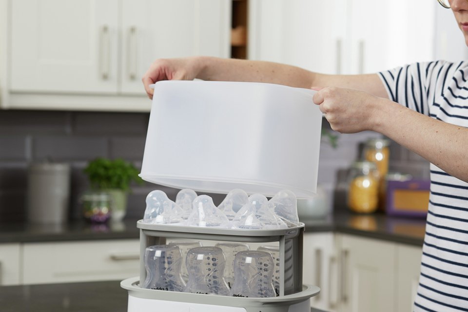 A man lifting the top off a Tommee Tippee super-steam advanced steriliser set with baby bottles in it.